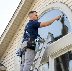 Professional window cleaner on a ladder using a squeegee to clean an exterior arched window, Fort Wayne, IN.